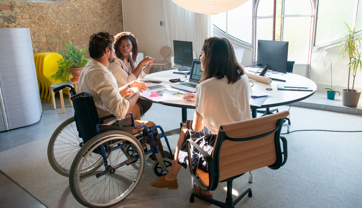 Un grupo de personas se reúne alrededor de una mesa en una reunión de trabajo, con uno de los participantes en silla de ruedas.