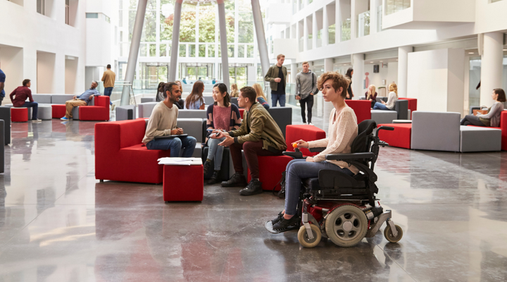 En un edificio con mucha iluminación, va una persona en silla de ruedas eléctrica. Parece una universidad. 